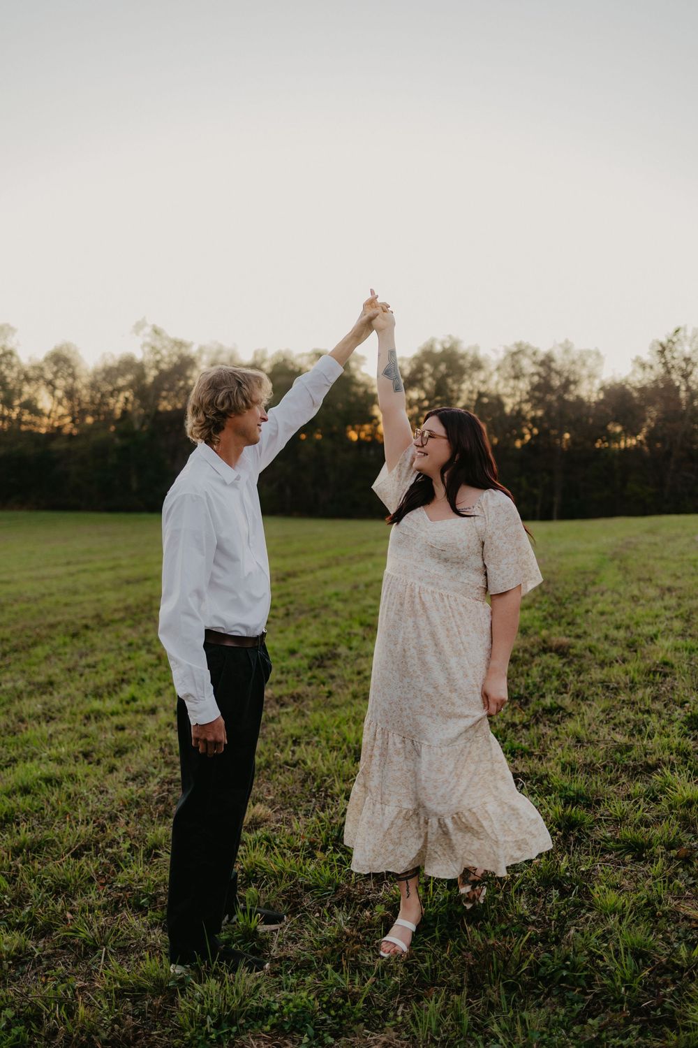Couple dancing in a green field at golden hour, man in white shirt and woman in floral dress twirling hands raised.