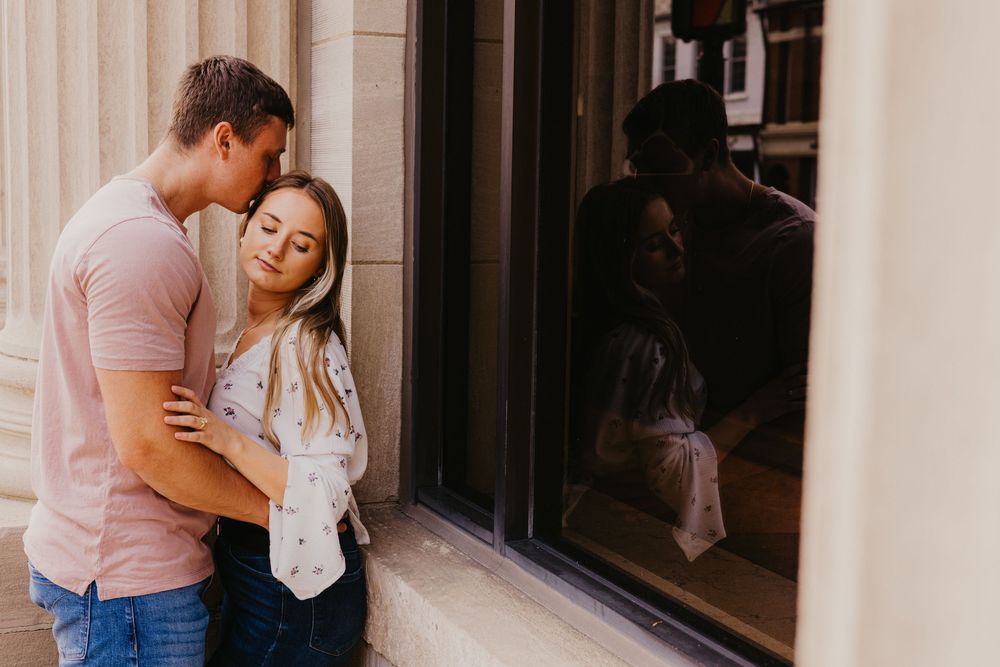 Romantic couple embracing tenderly near a window, man kissing woman's head in a warm outdoor setting.