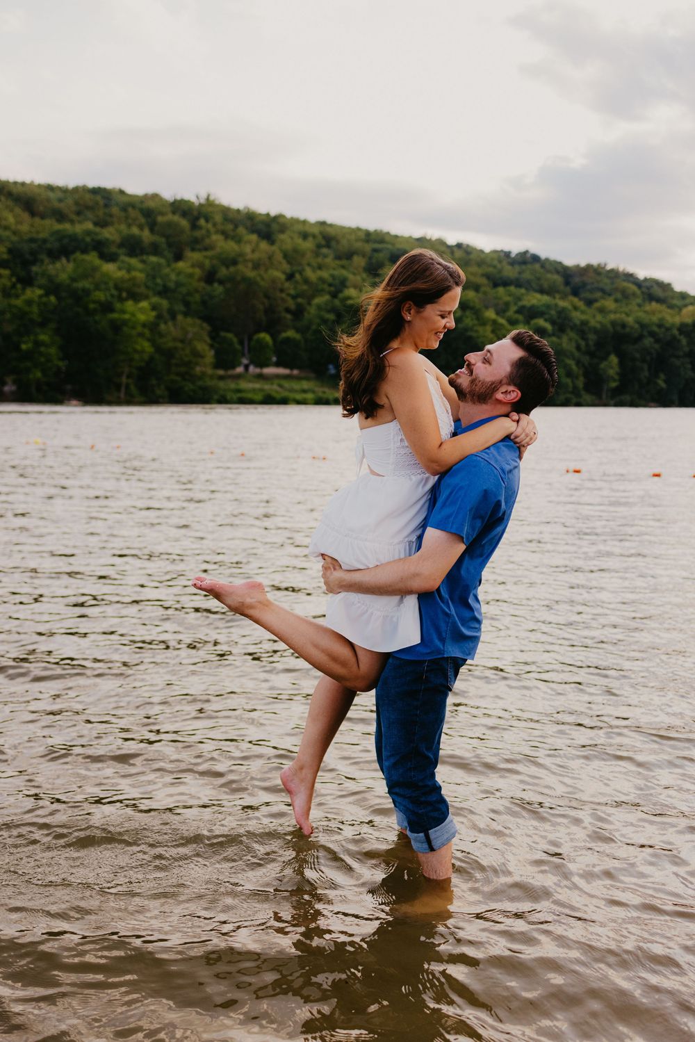 A man lifts a woman in his arms while standing in a lake, sharing a romantic moment with lush green hills behind.