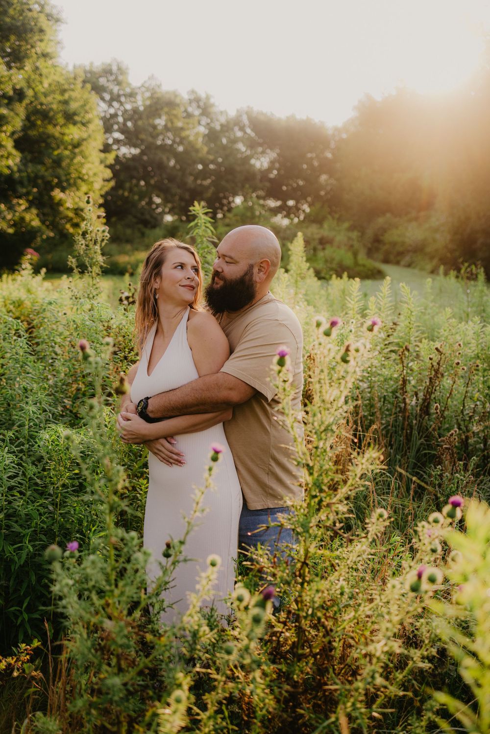 Couple embracing in a sunlit wildflower field, man with beard hugging woman in white dress during golden hour.