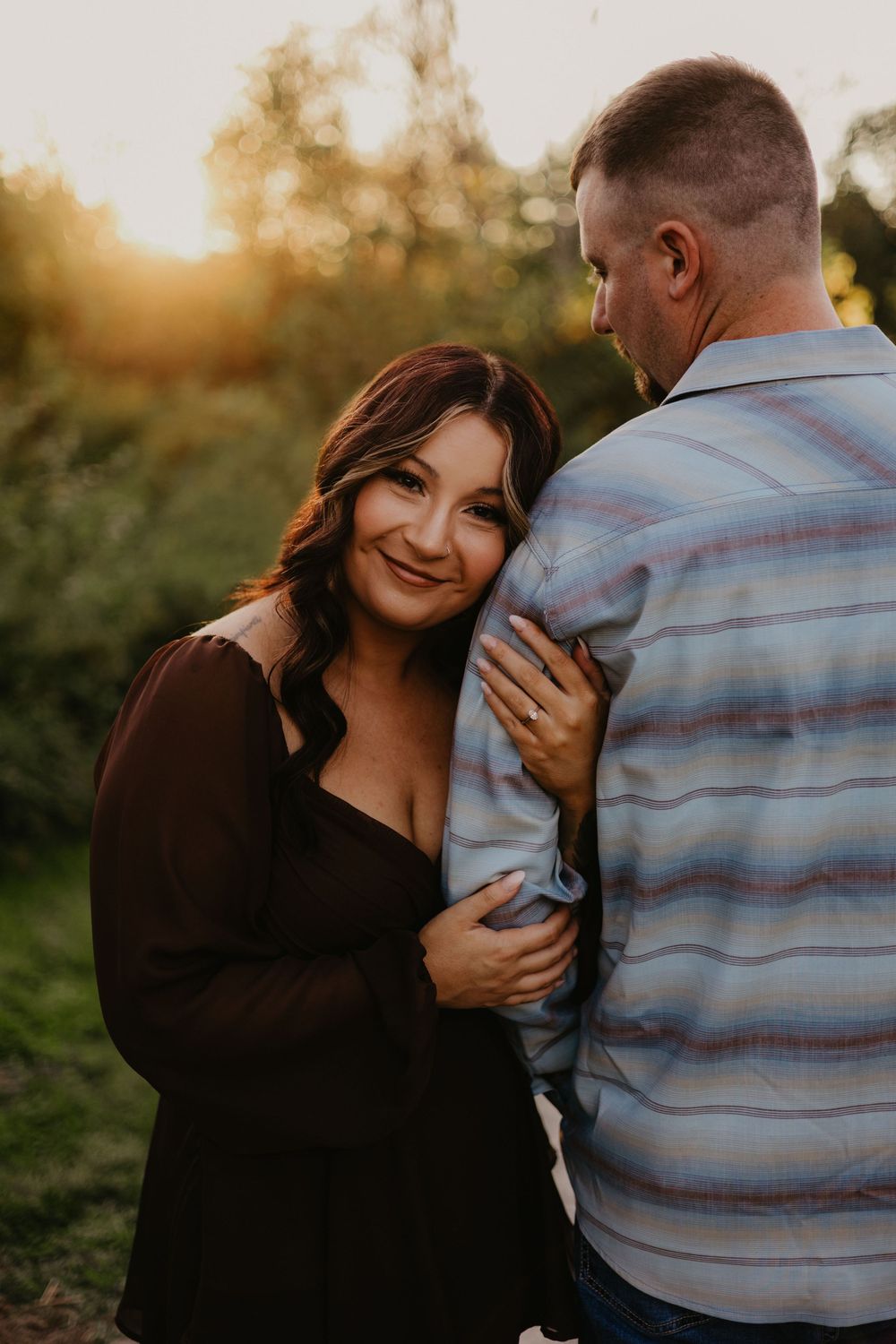 A woman in a black dress smiles while embracing a man in a striped shirt during a golden hour outdoor photo session.