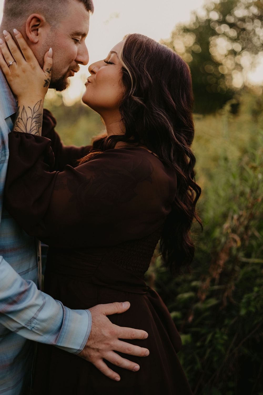 A couple shares a romantic kiss outdoors at golden hour, surrounded by lush greenery, as she wears a dark brown dress.