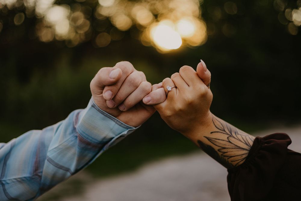 Close-up of couple doing a pinky promise, one partner showing a tattoo sleeve and engagement ring.