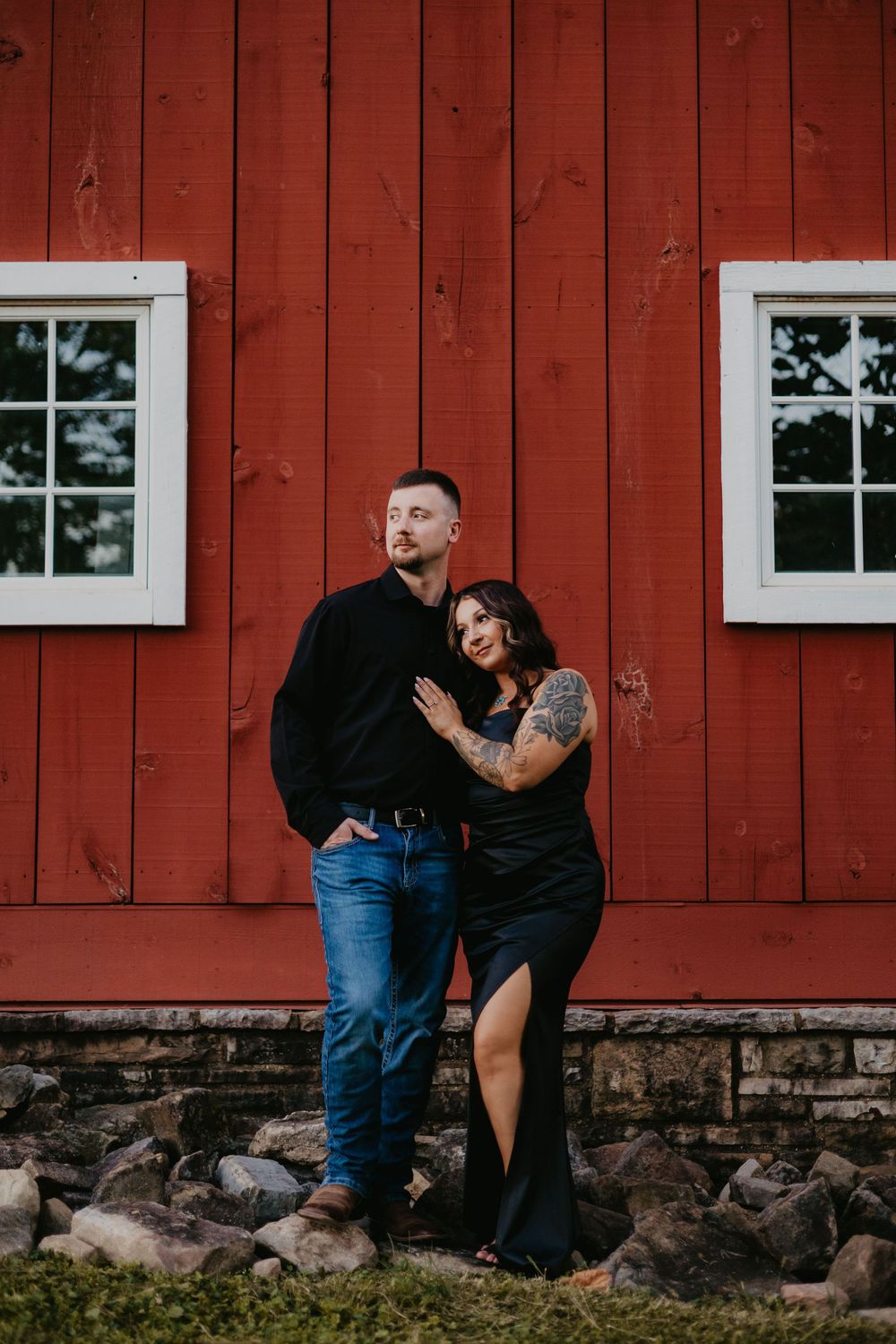 Couple poses against a rustic red barn wall, woman with tattoo in black dress leaning on partner.