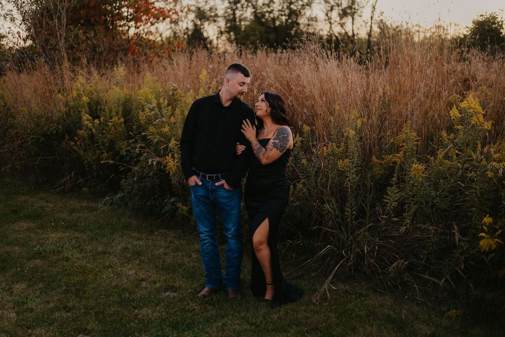 Couple posing together at golden hour in a field of tall grasses, woman in black dress with tattoos.
