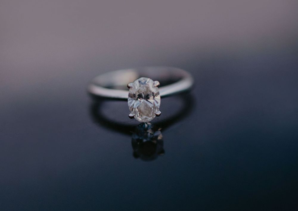 Elegant solitaire diamond engagement ring with oval cut stone set in silver band on dark reflective surface.