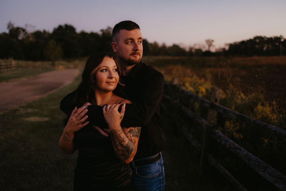 Couple embracing at sunset near a wooden fence in an open field, man hugging woman from behind.
