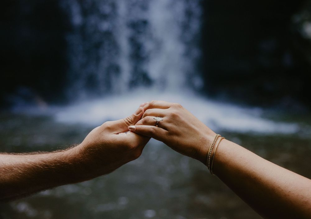 Two people holding hands tenderly in front of a waterfall, one wearing a gold bracelet.