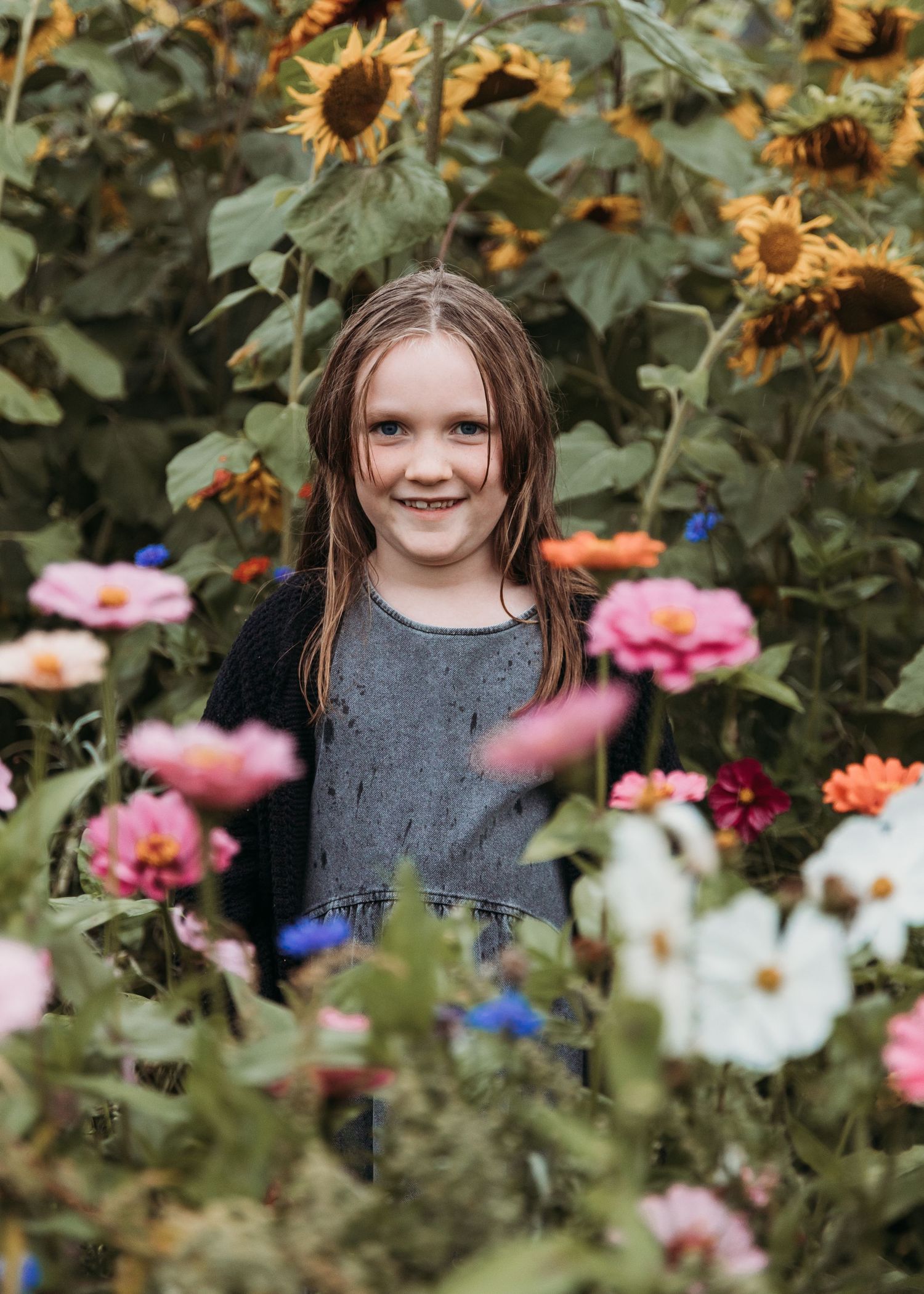A smiling young girl with braids stands among colorful zinnias and sunflowers in a lush garden.