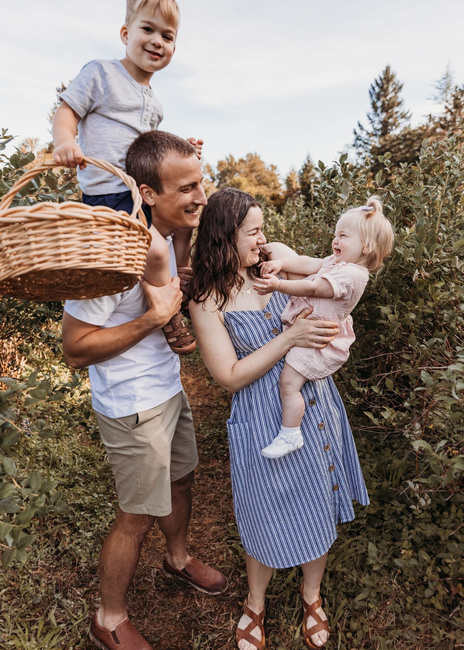 A happy family of four picks berries outdoors, with dad holding a basket and toddler while mom carries a baby girl.