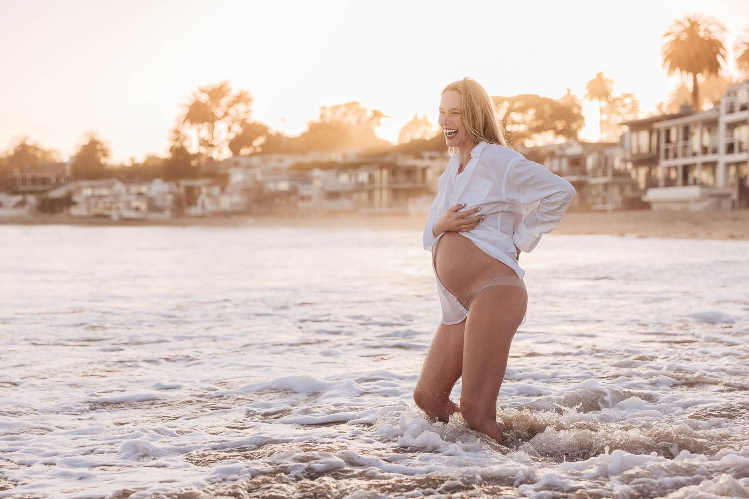 A person in a white shirt and bikini bottom plays on a sunny beach at sunset with coastal homes in the background.