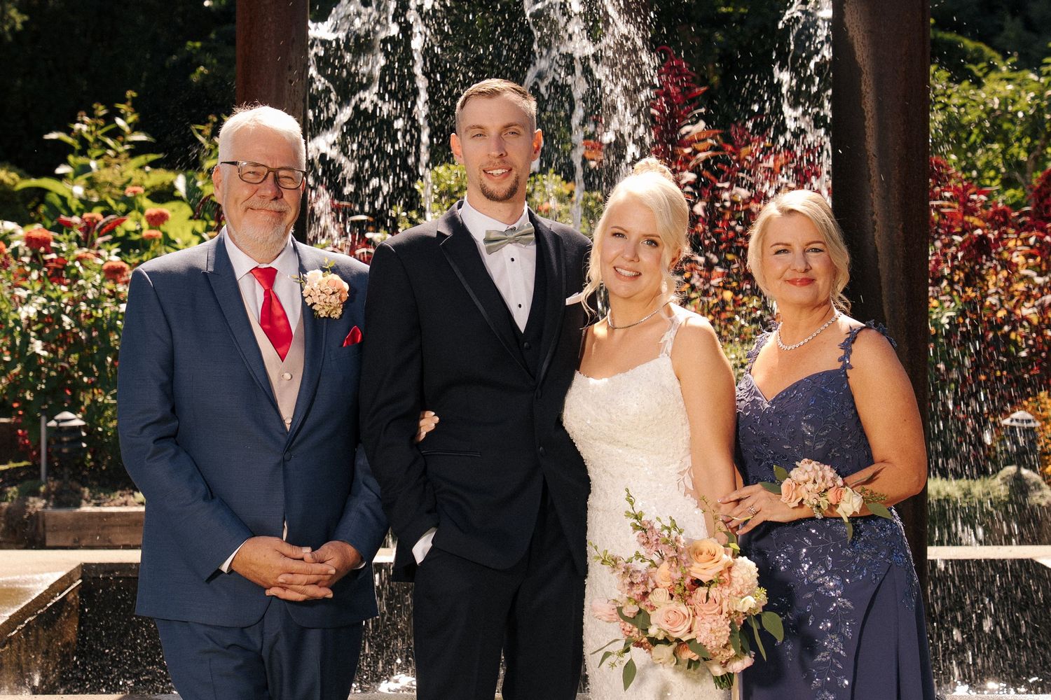 Wedding party of four posing in front of a fountain, bride in white gown holding bouquet, groom in tuxedo, family in formal attire.