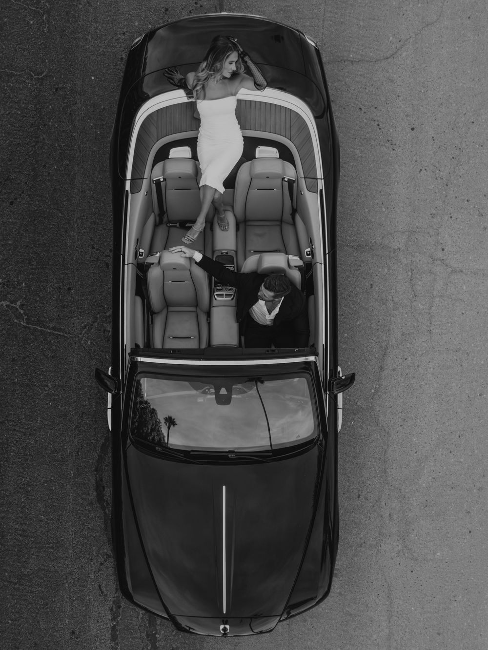 Aerial black and white photo of a convertible car with a woman in white dress posing on the hood.