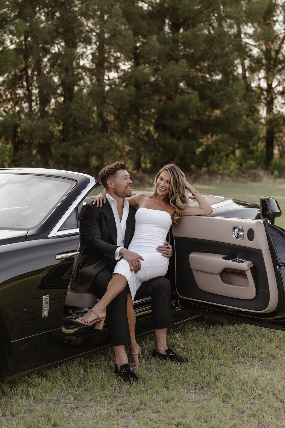 Elegant couple in formal attire posing beside a luxury convertible car in a scenic outdoor setting.