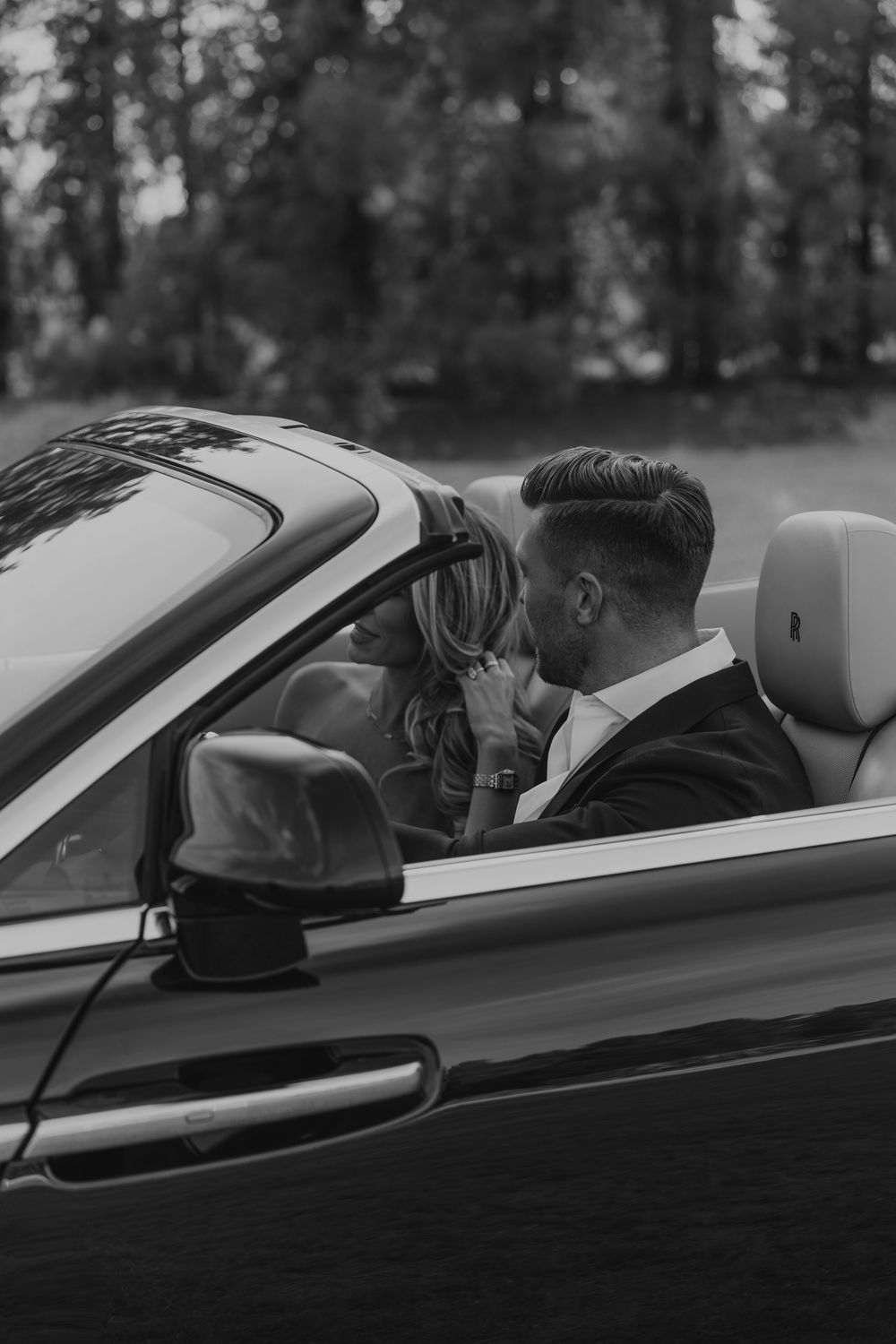 Black and white photo of a couple sharing a romantic moment in a luxury convertible car surrounded by trees.
