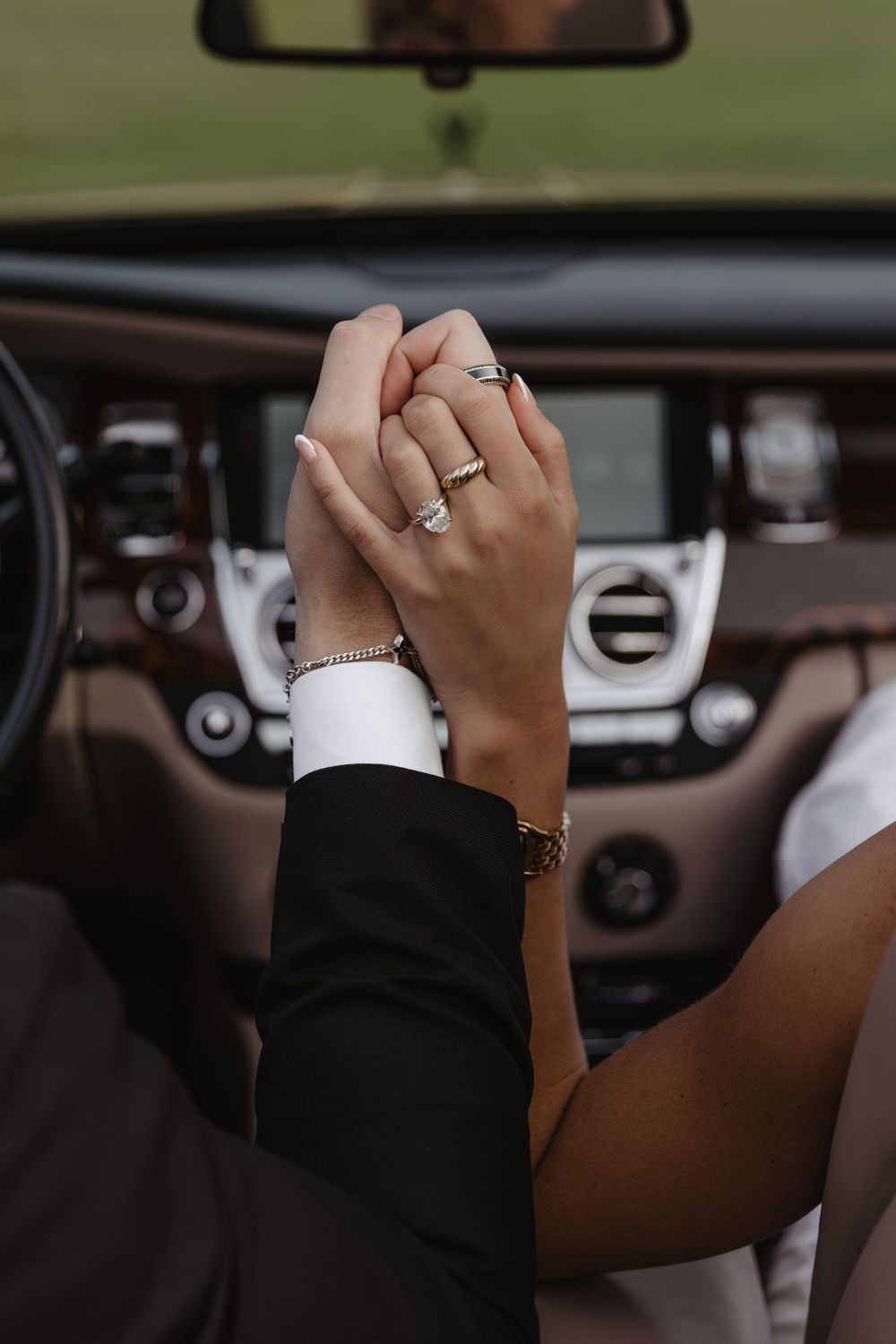 Close-up of couple holding hands showing wedding and engagement rings inside a luxury car interior.