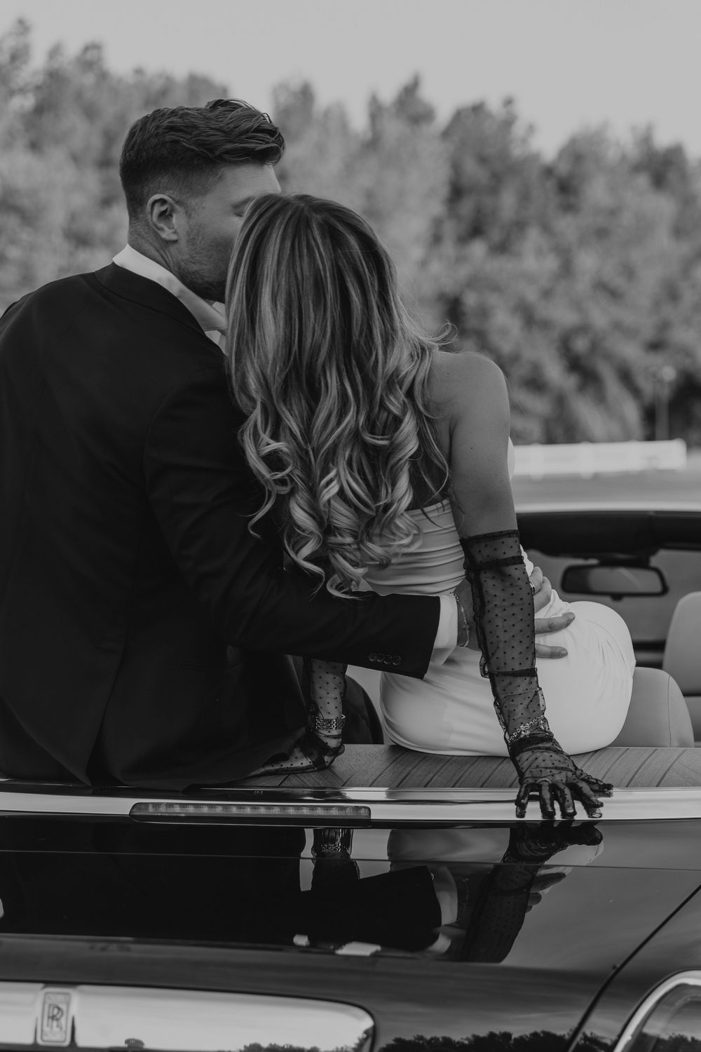 Black and white photo of a couple sharing a romantic kiss while sitting on a classic convertible car outdoors.