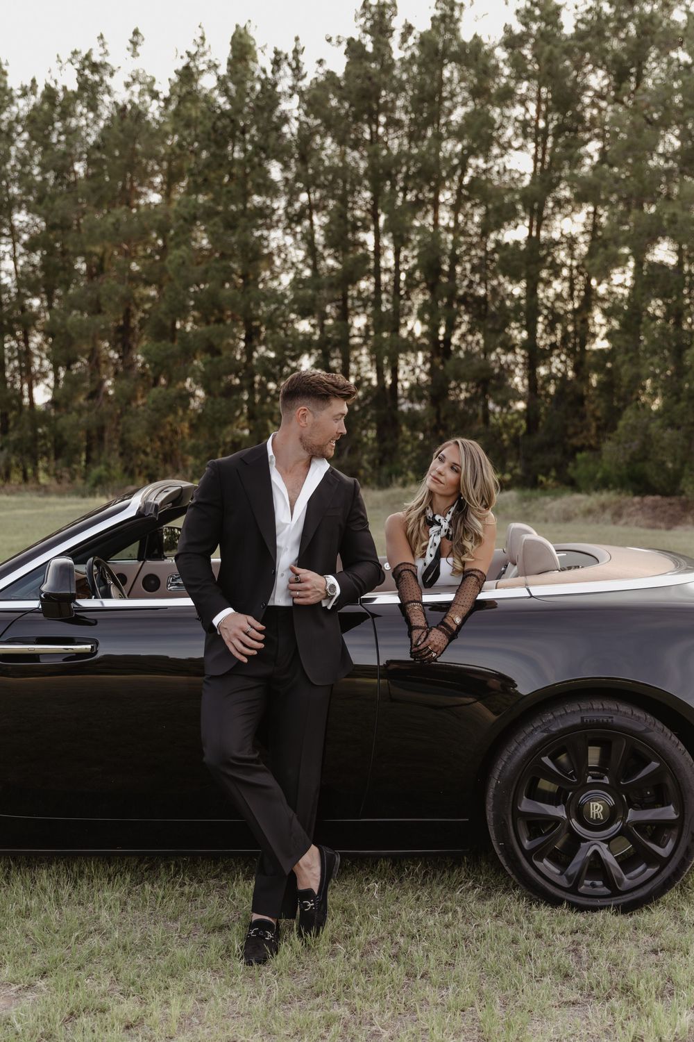 Elegant couple posing beside a black Rolls-Royce convertible in a scenic outdoor setting with pine trees.