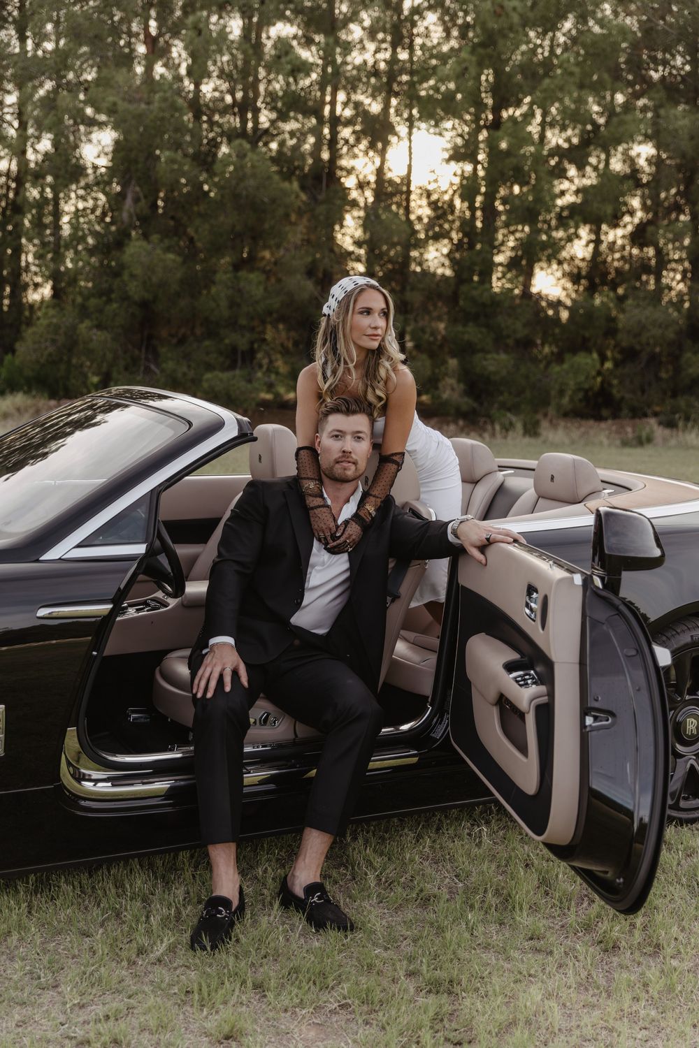 Stylish couple in wedding attire posing with a black luxury convertible car at sunset outdoors.