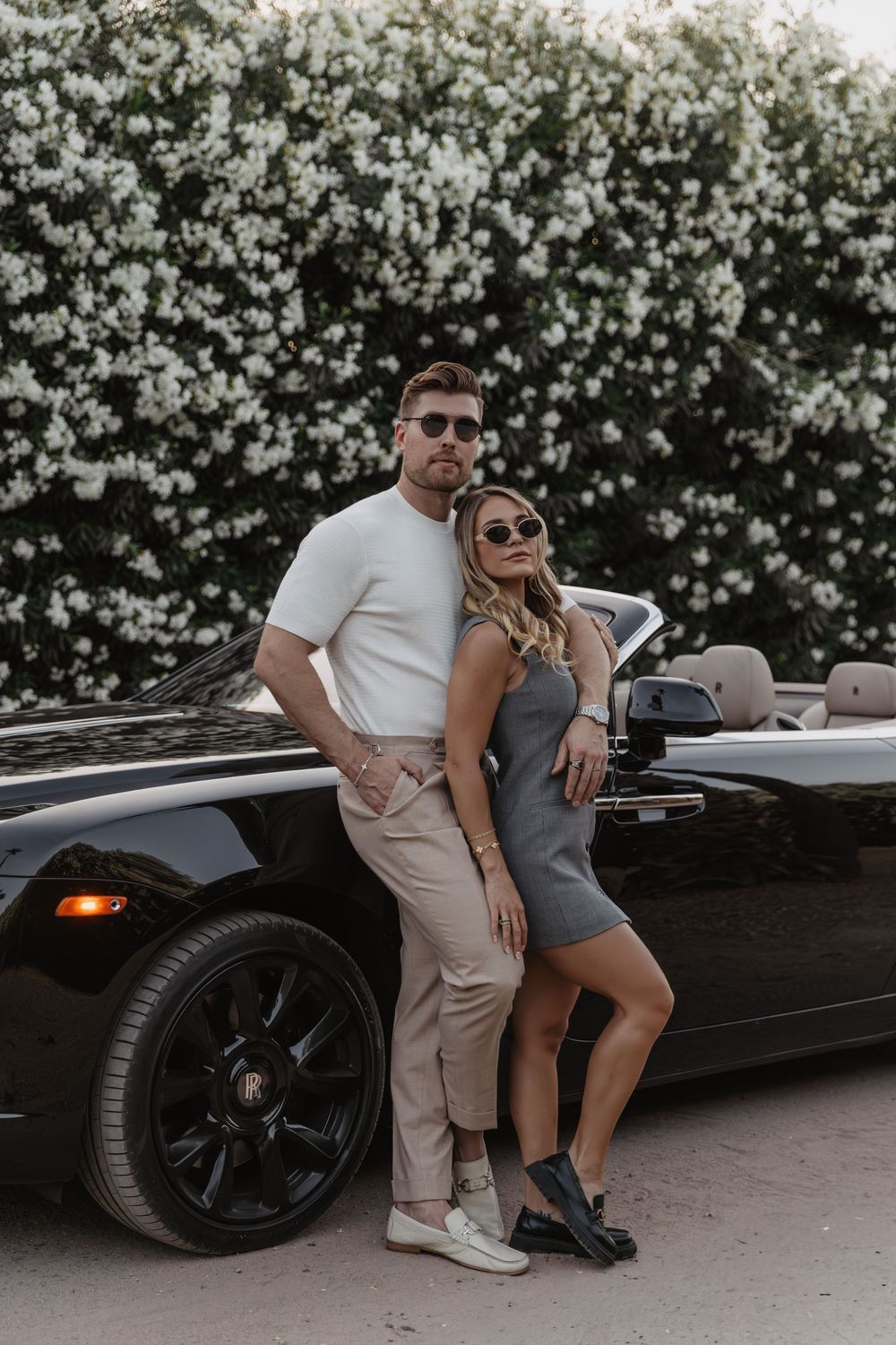 Stylish couple posing in front of a black convertible Porsche with white flowering hedges in the background.