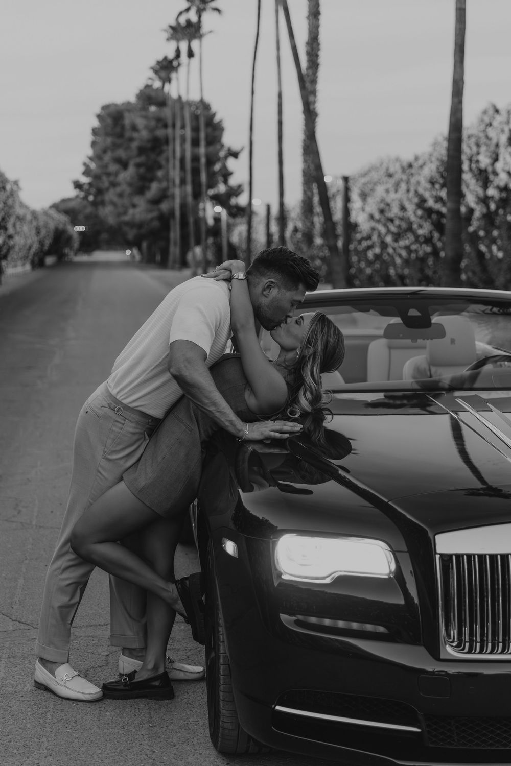 Romantic couple kissing beside a luxury Rolls-Royce convertible on a palm-lined street.