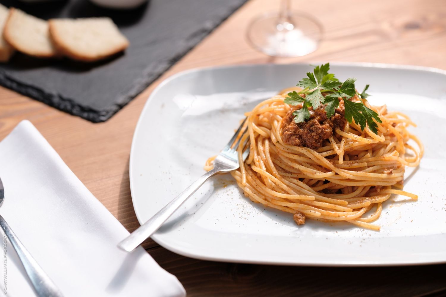 A plate of spaghetti bolognese topped with fresh parsley, served on a white plate with bread and wine in the background.