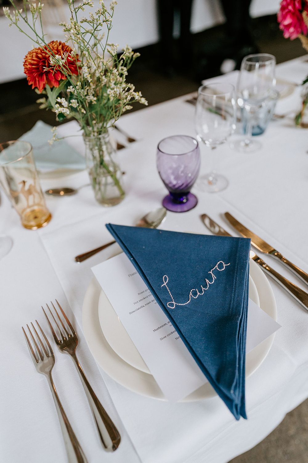 Elegant wedding place setting with blue folded napkin embroidered with Laura, gold cutlery, and colorful glassware.
