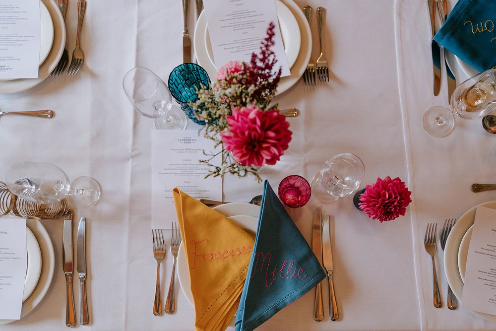 Elegant wedding table setting with colorful teal and gold napkins, pink dahlias, white plates, and gold cutlery.