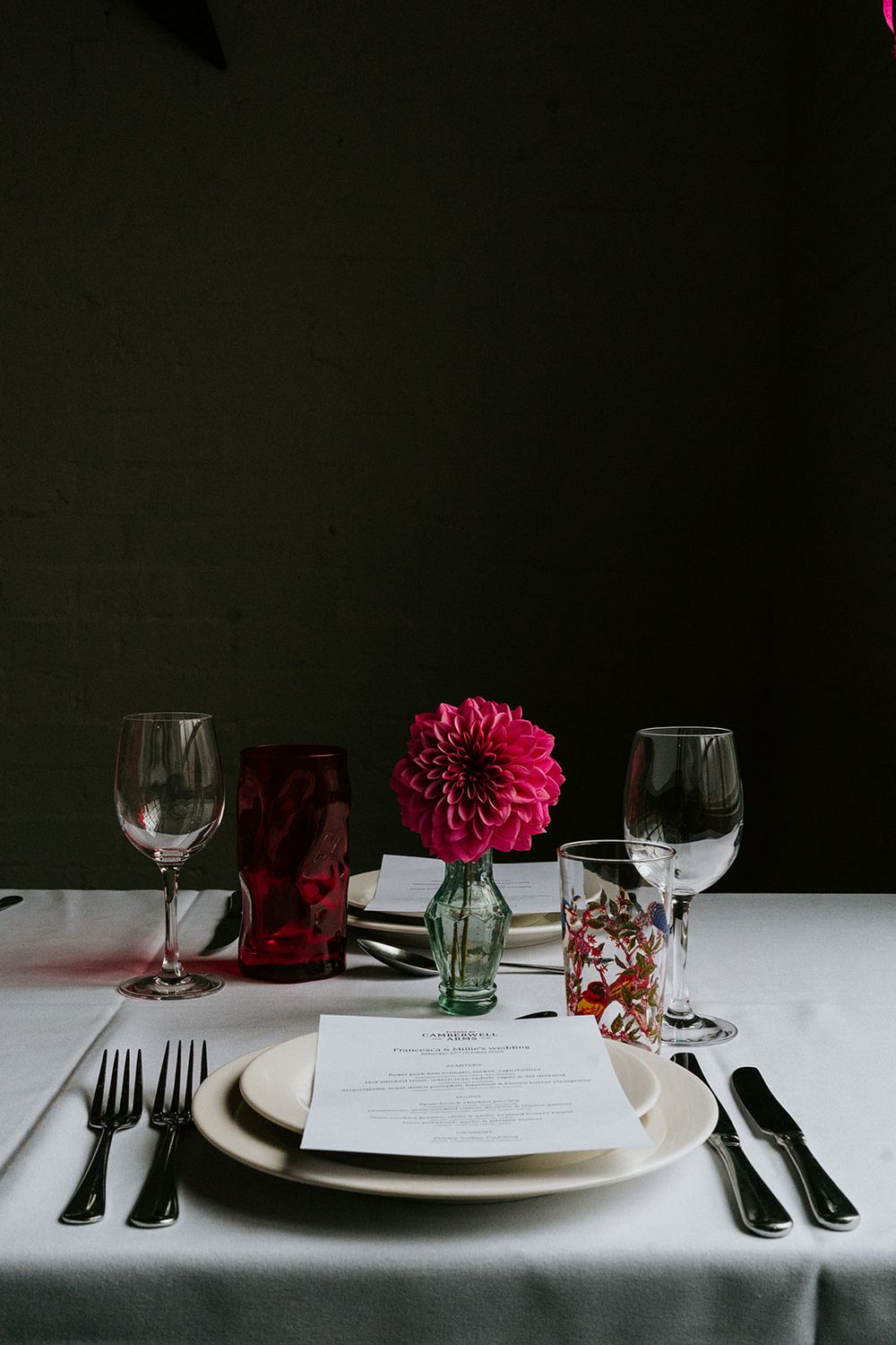 Elegant table setting with pink dahlia centerpiece, wine glasses, and menu card on white linen.