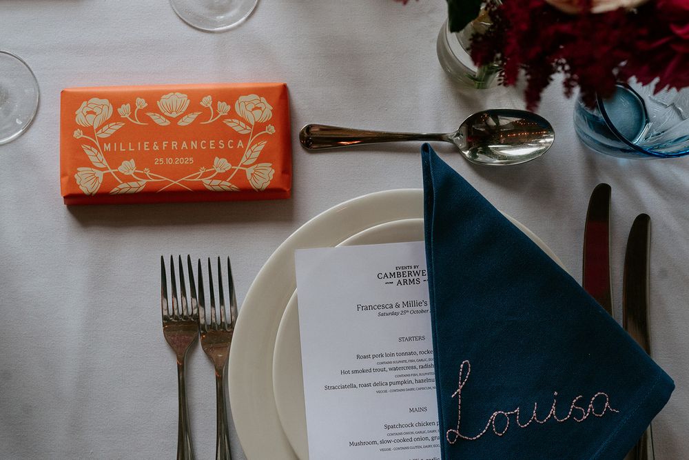 Elegant wedding place setting with blue napkin, menu card, silverware, and orange personalized chocolate favor box.
