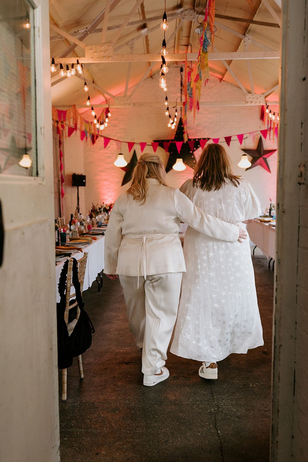 Two brides in white outfits walk arm-in-arm into a festively decorated wedding reception venue with colorful bunting.