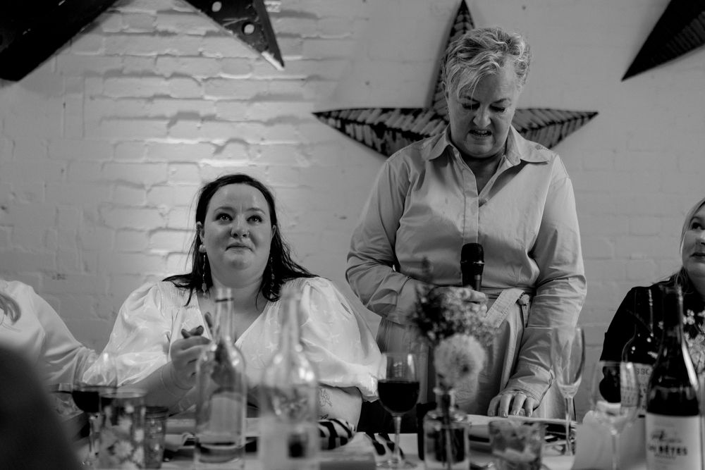Black and white photo of two women at a wedding reception dinner table, one seated smiling, one standing speaking.