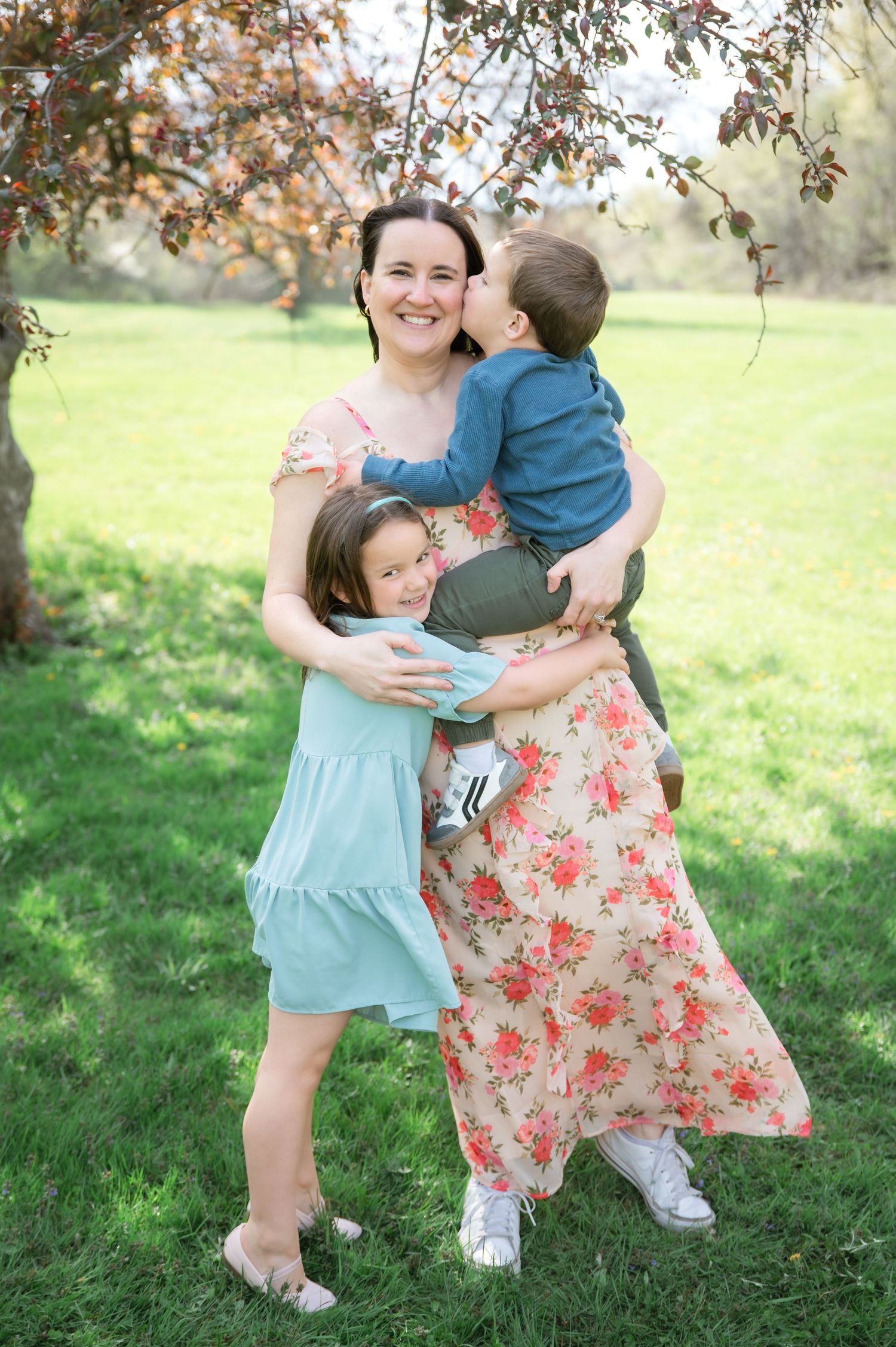 A smiling woman in a floral dress holds two young children in a sunny park, sharing hugs and kisses outdoors.