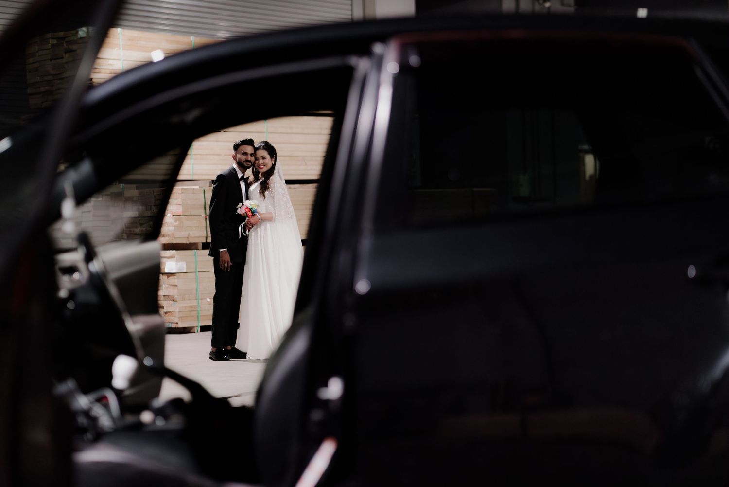 A couple in wedding attire stands under a fluorescent light, the groom in a suit and bride in a white hijab and lace gown.