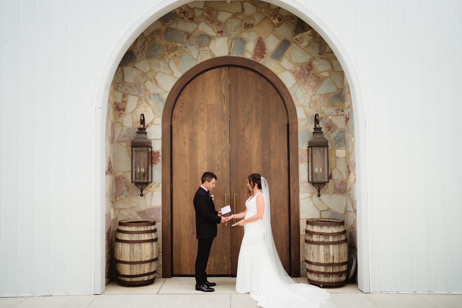 Bride and groom exchange vows before grand wooden arched doors with stone accents and barrel decor at elegant wedding venue.