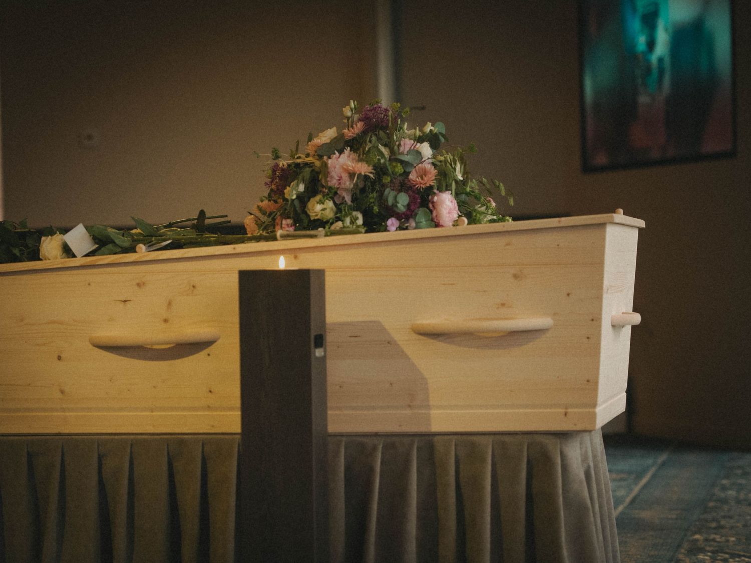 A wooden coffin adorned with pink and purple flowers rests on a draped stand in a funeral home.
