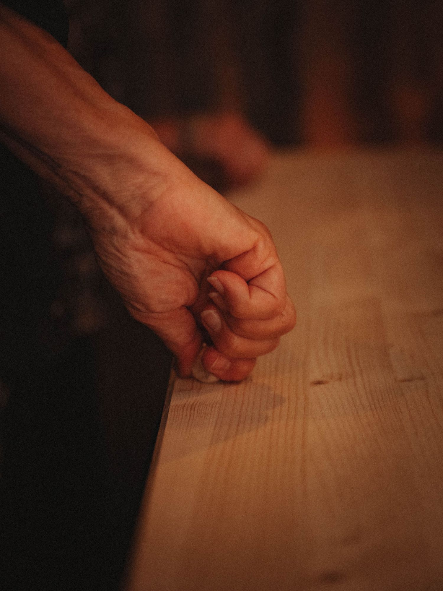 A clenched fist resting on a wooden table surface in dramatic moody lighting.