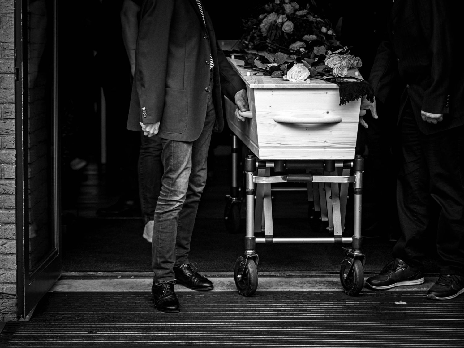 Black and white photo of pallbearers carrying a white casket with flowers at a funeral service.