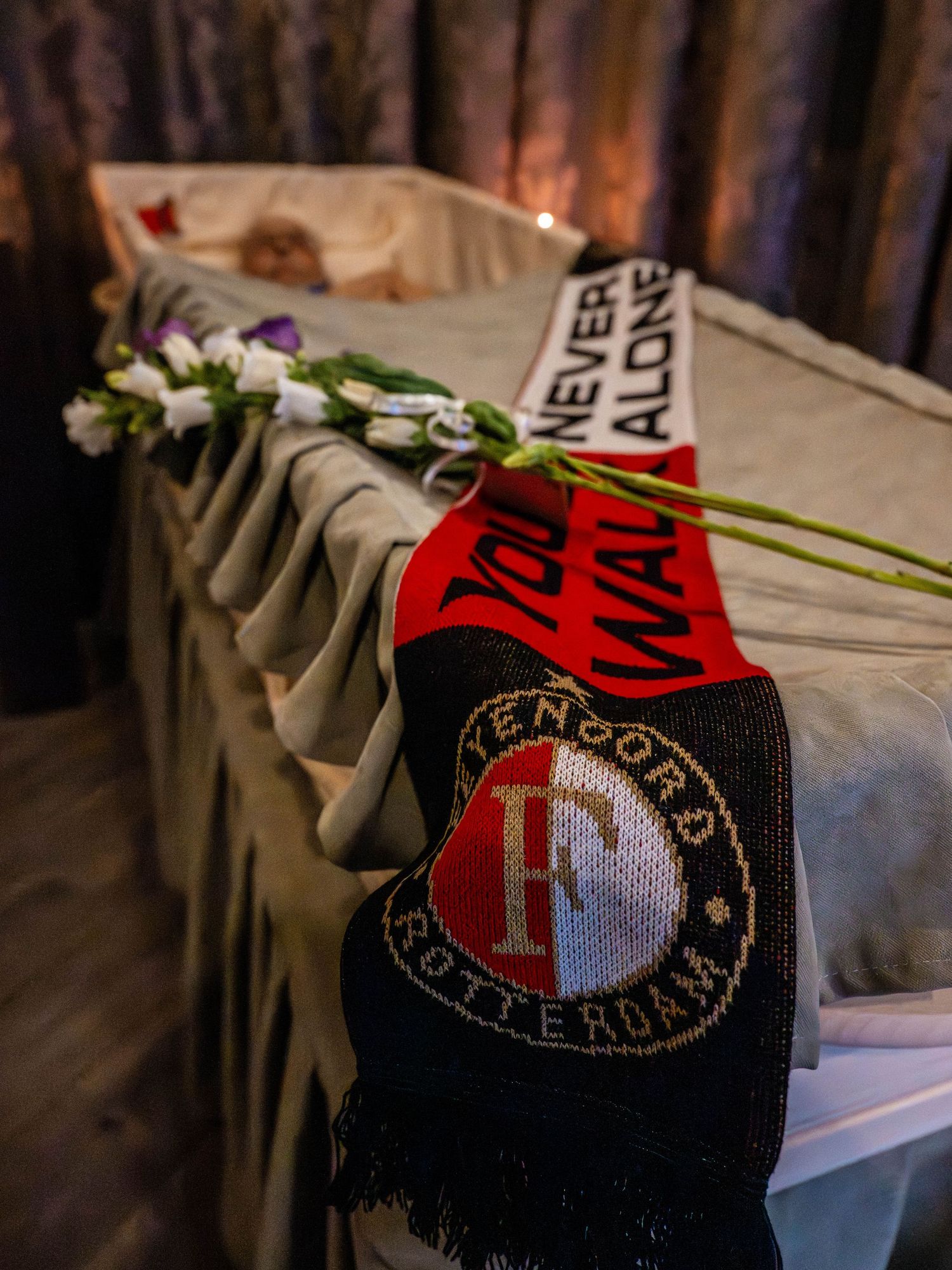 Feyenoord Rotterdam scarf draped over a casket with white flowers and You'll Never Walk Alone text at a funeral.