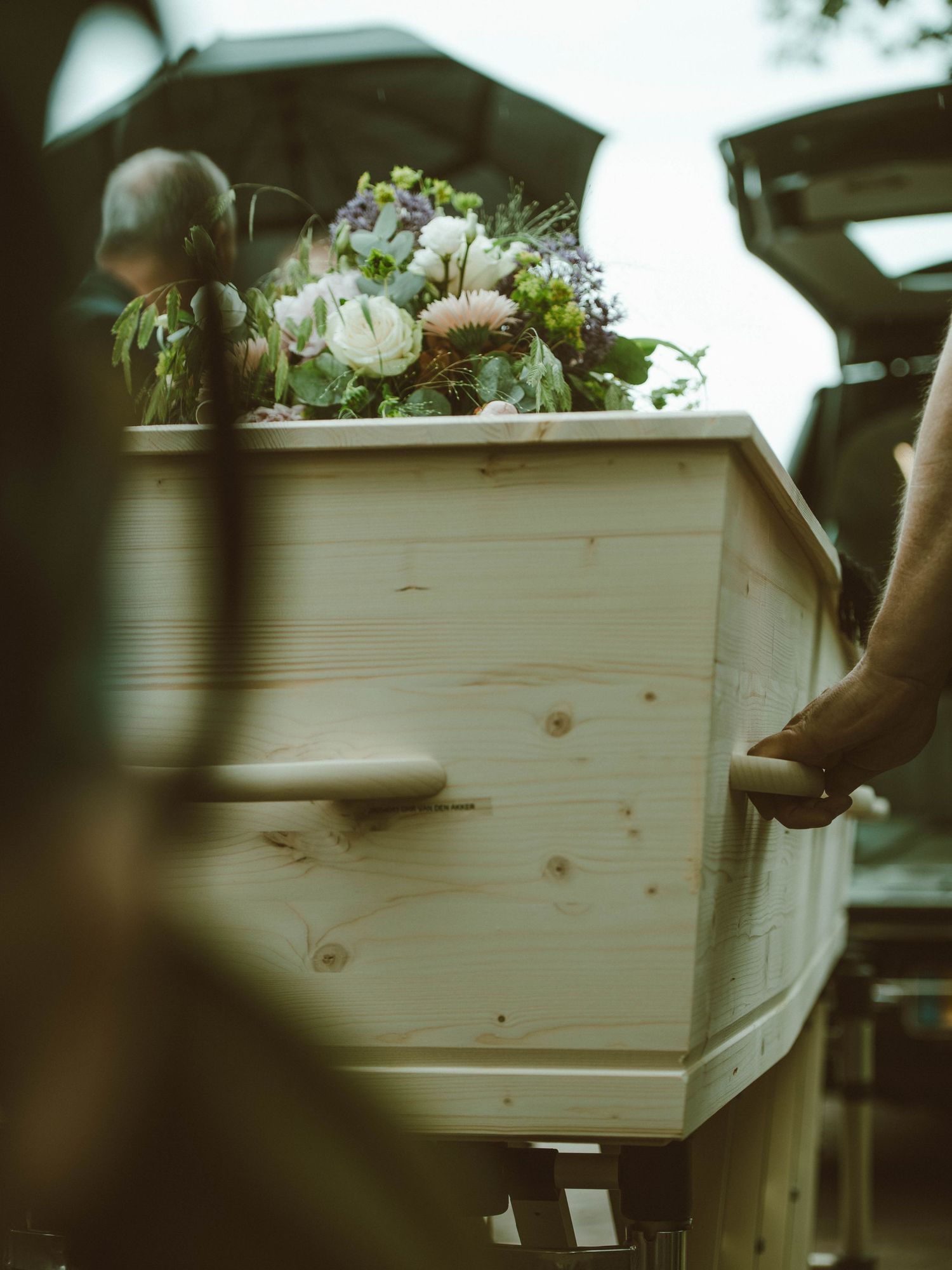 A light wood coffin adorned with flowers being carried at a funeral service outdoors.