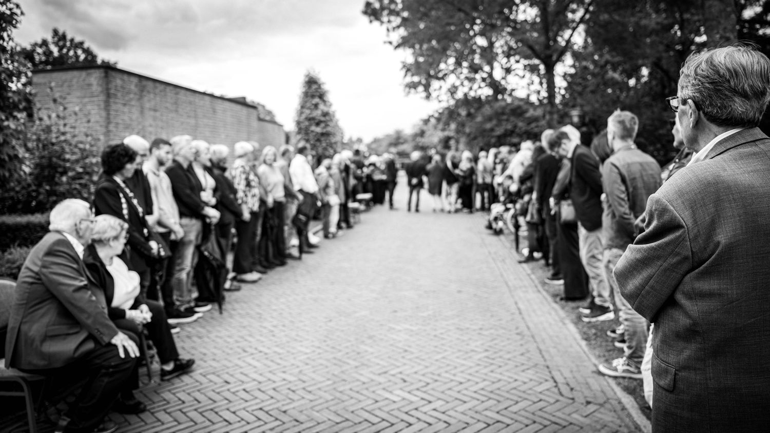 Black and white photo of a large group of people standing in a line along a brick path outdoors.