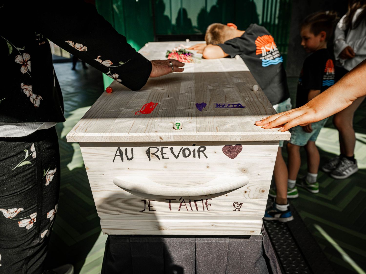 White wooden box with French writing Au Revoir Je t'aime surrounded by people touching it at an event.