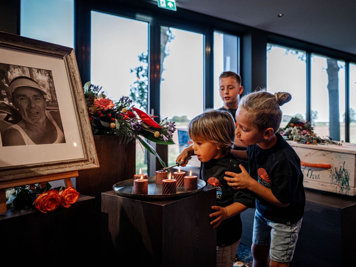 Children hugging at a memorial service with candles and a framed photo surrounded by flowers.