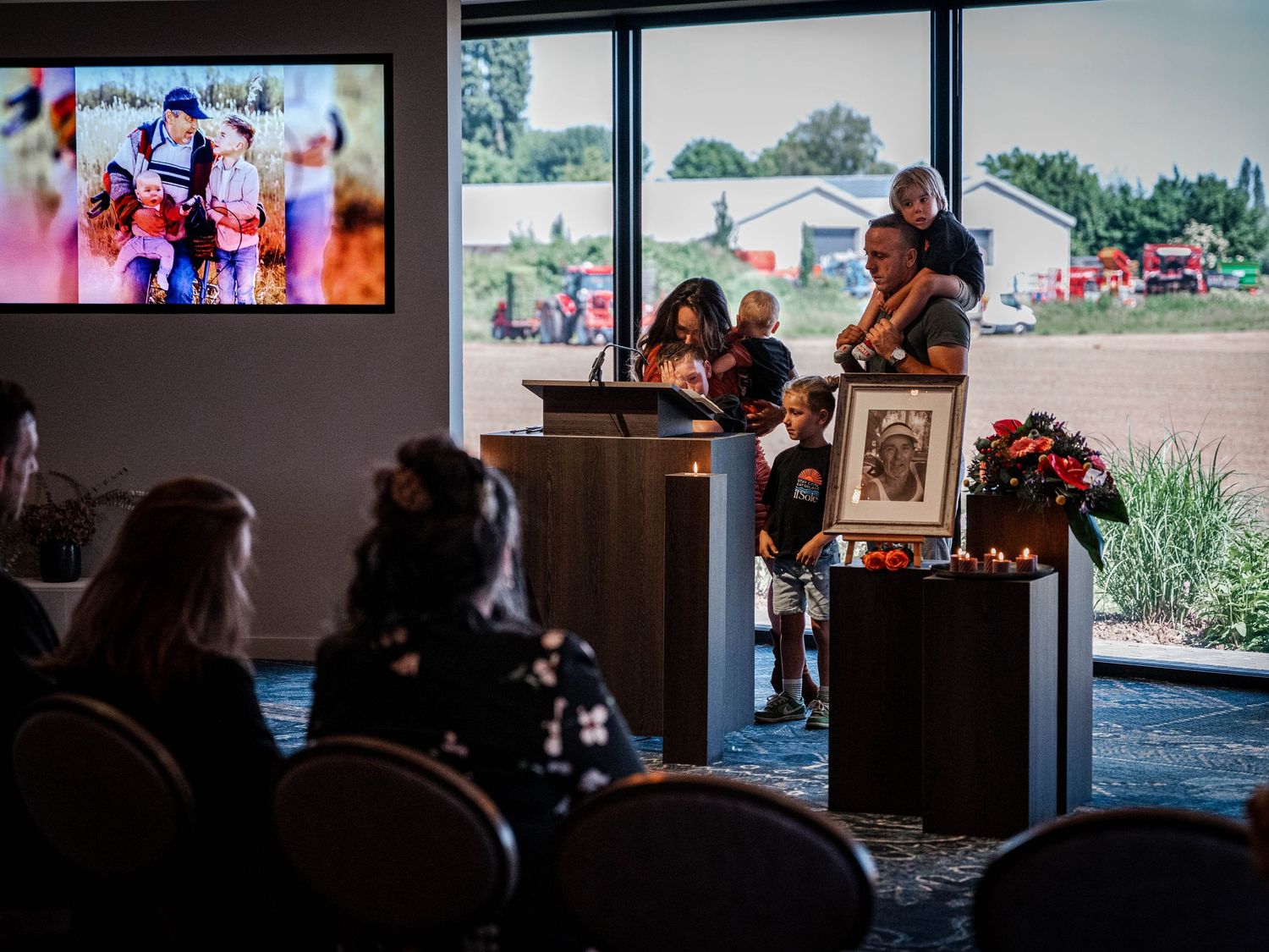 A family gathers at a memorial service podium with a framed portrait and floral arrangement, as a photo displays on screen.