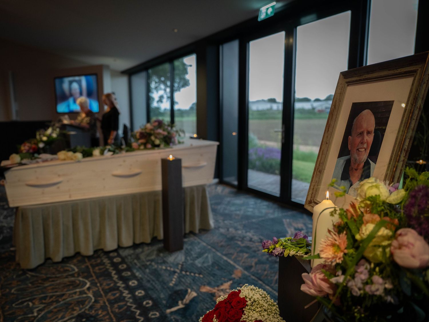 Funeral home interior with white casket, floral arrangements, framed portrait, and candles in a softly lit memorial room.