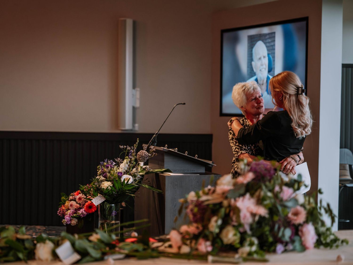 Two people embrace at a memorial service with flowers in foreground and a portrait displayed on screen behind them.