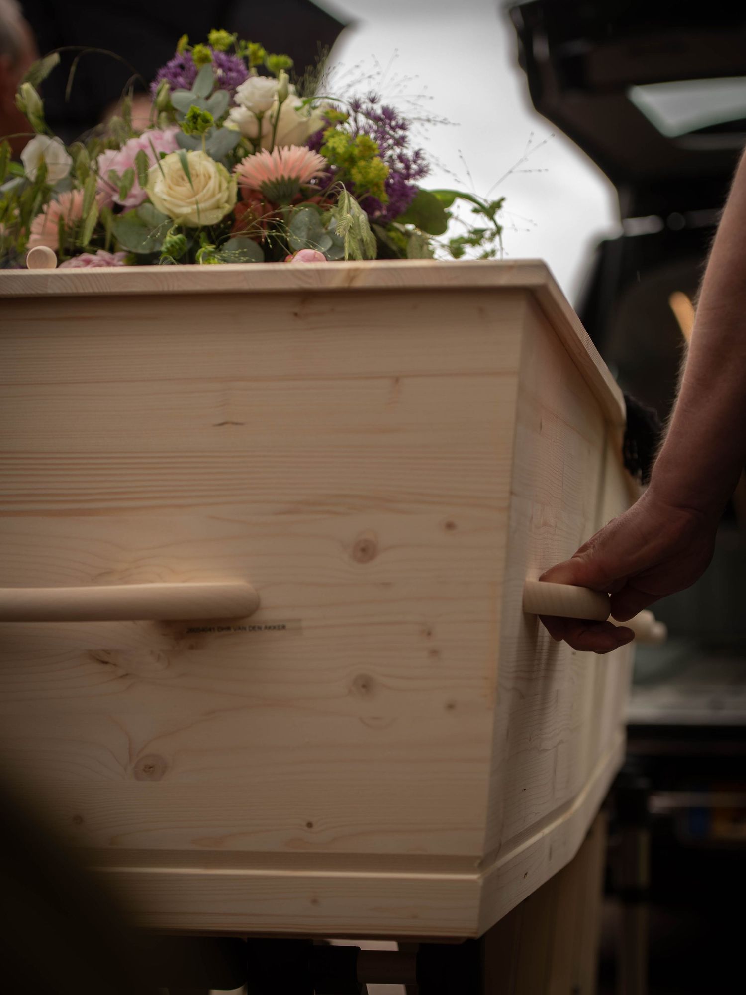 A light wood coffin adorned with colorful flowers being carried by pallbearers at a funeral service.