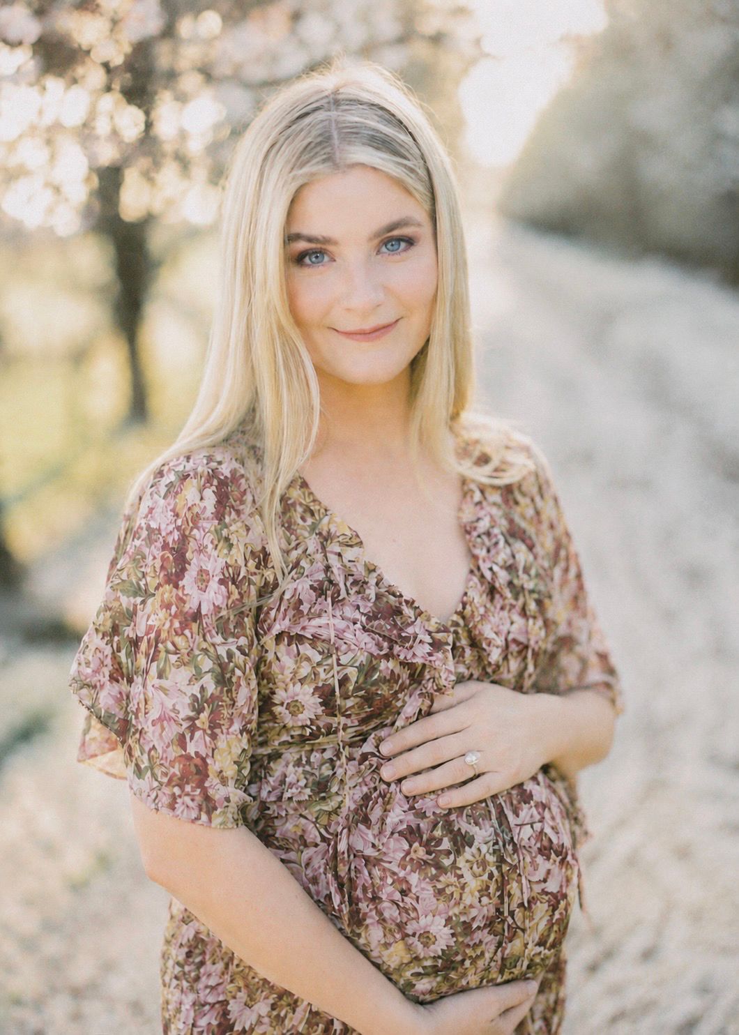 Pregnant woman in floral dress cradling baby bump outdoors during golden hour maternity photo session.