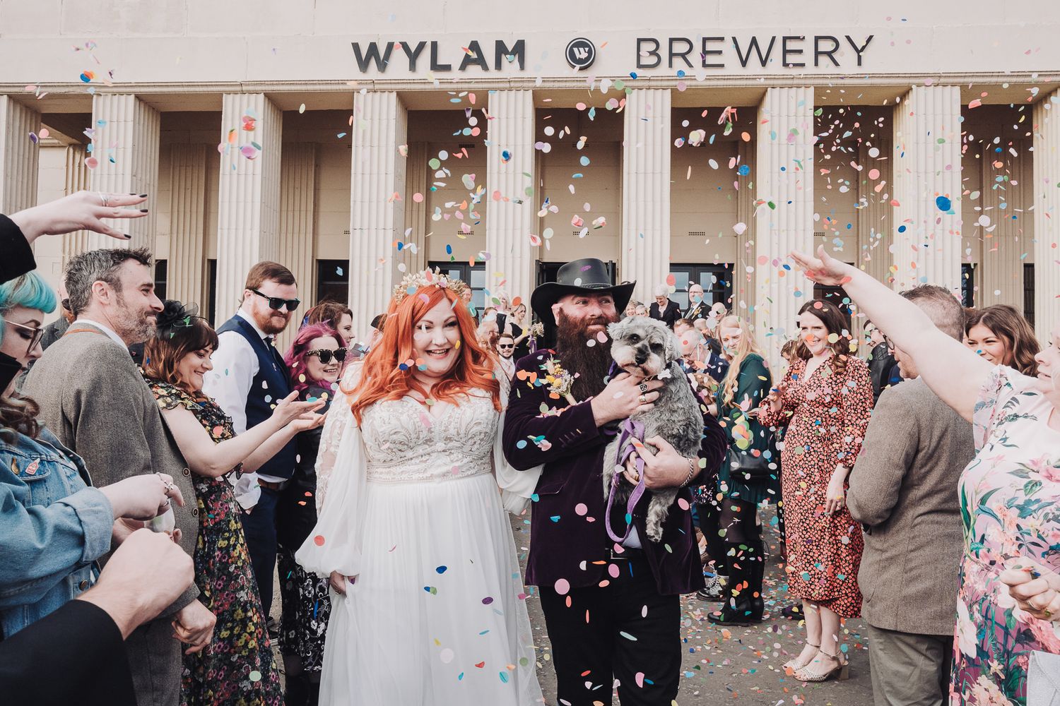 Bride and groom walk through confetti shower outside Wylam Brewery wedding venue, surrounded by cheering guests.