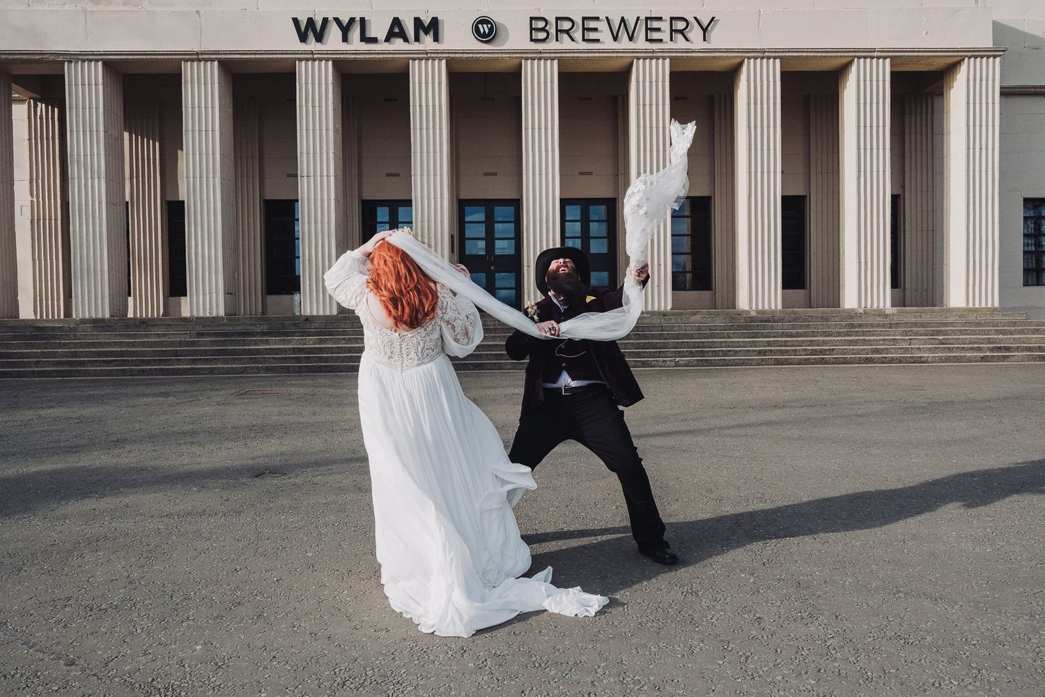 Bride and groom dancing playfully outside Wylam Brewery, bride in white gown with red hair, groom in black suit.